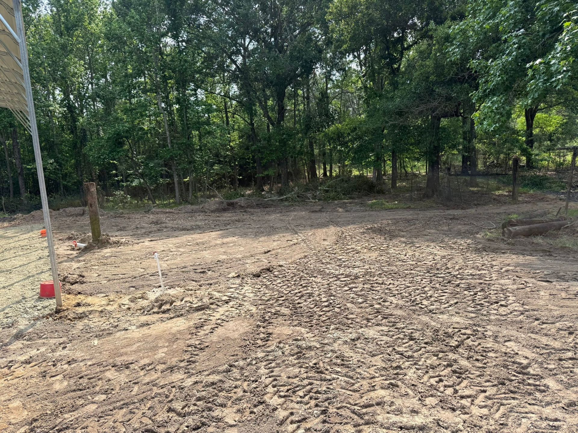 Bare dirt clearing beside a wooded tree line, with a red-and-white pole at the left edge.
