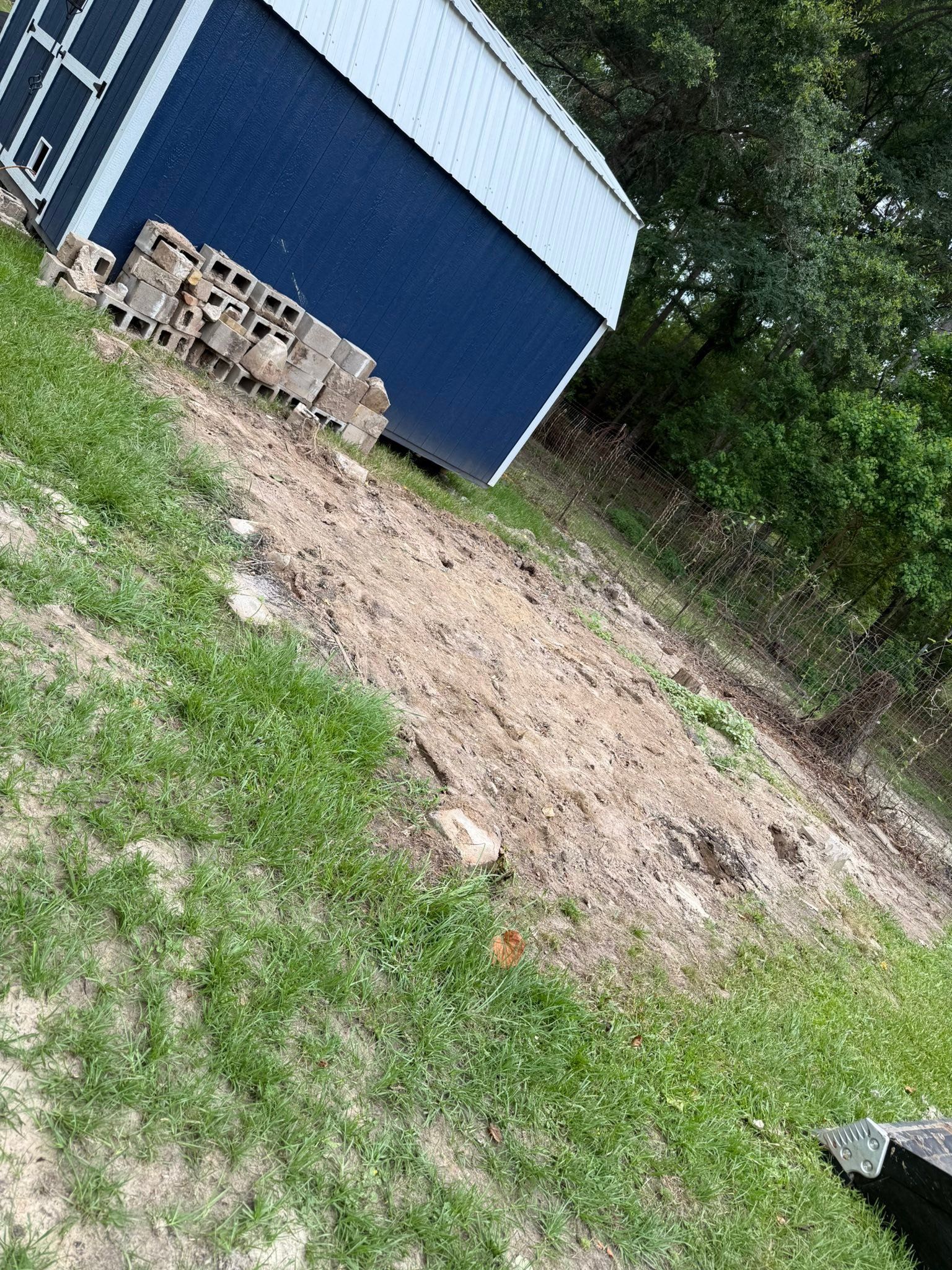 Blue shed beside a dirt patch and grassy yard, with stacked bricks near the wall.