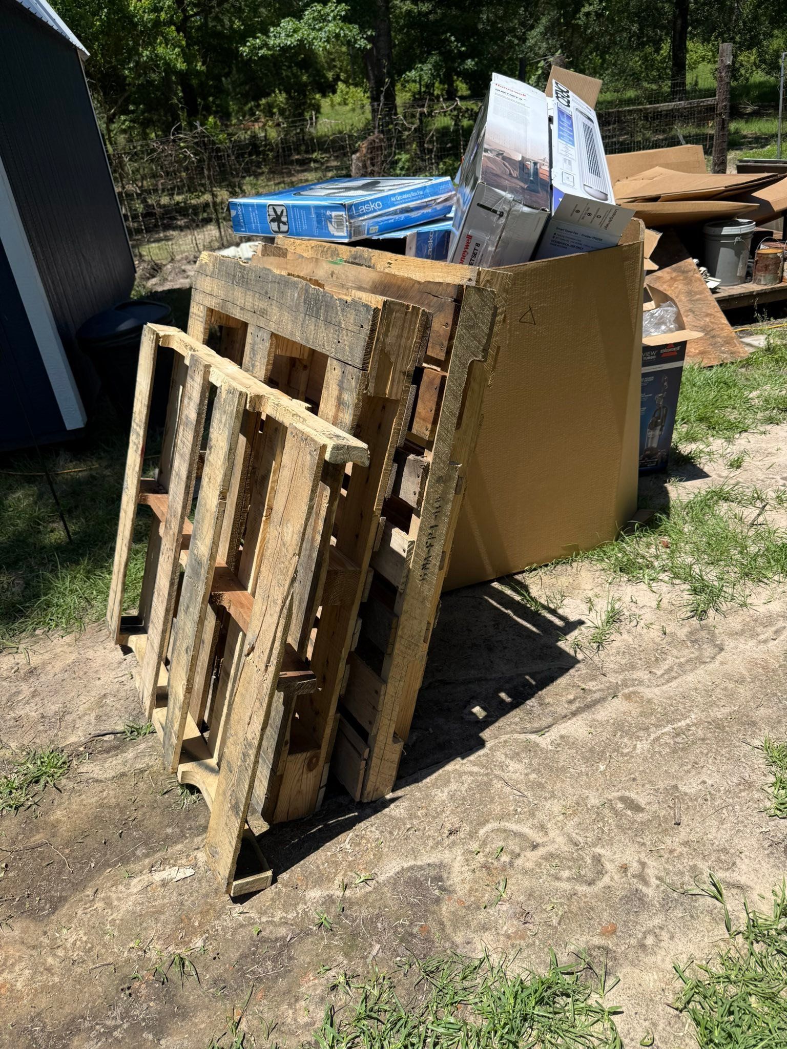 Wooden pallets and cardboard stacked outdoors on dirt beside a blue container.