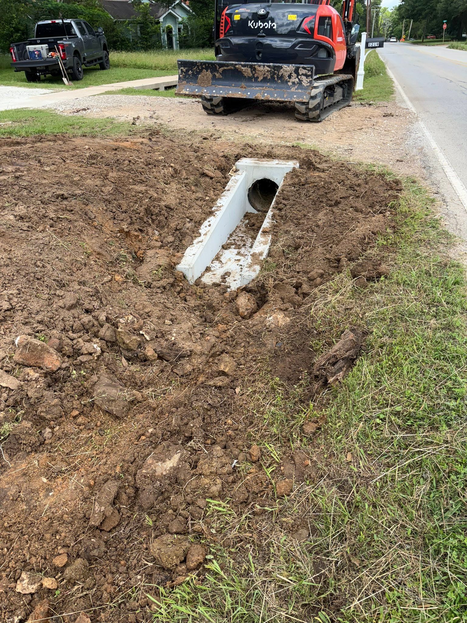 Damaged roadside culvert pipe exposed in muddy ditch beside a road and a tow truck