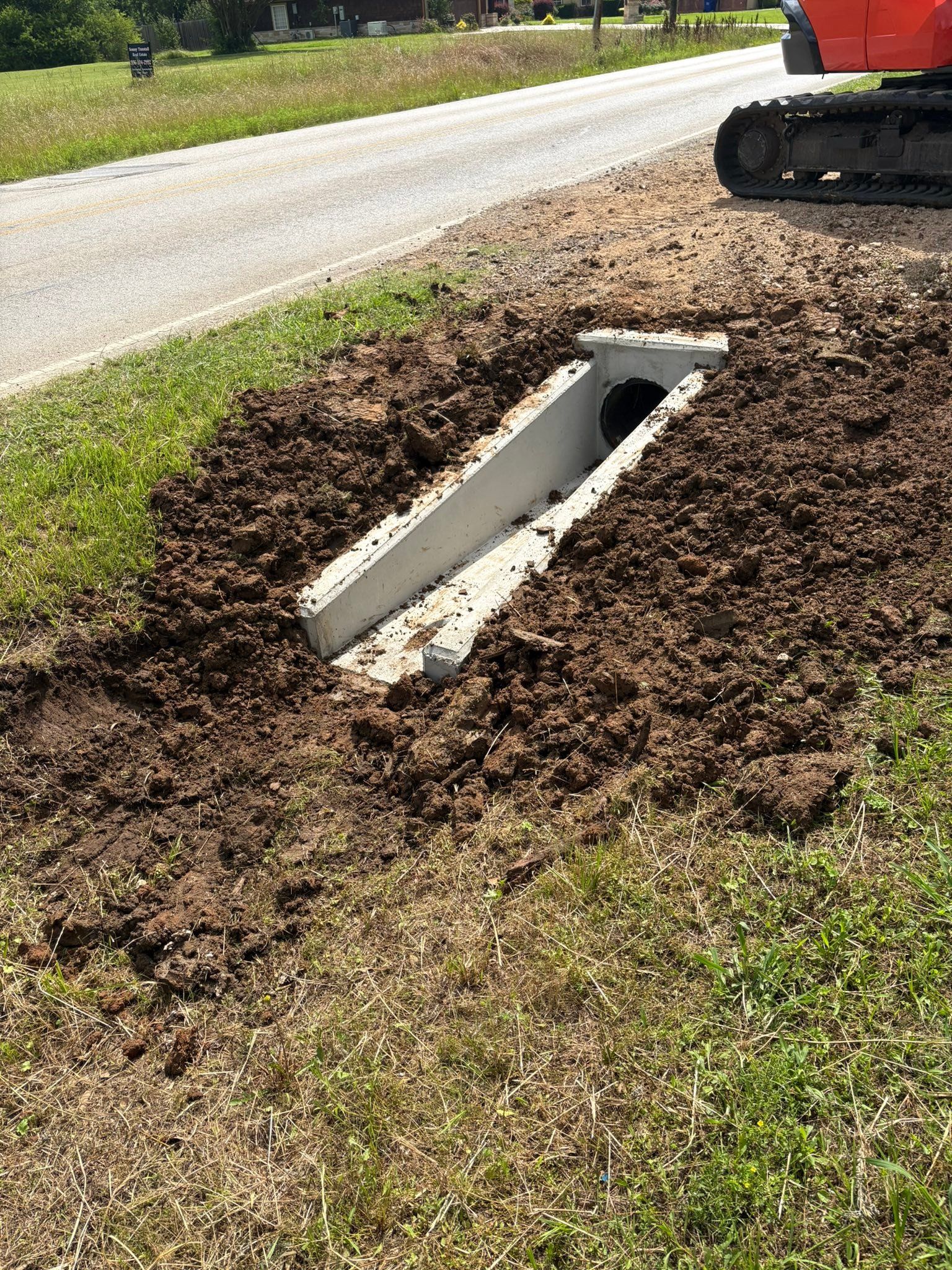 A narrow concrete drainage culvert in a dirt ditch beside a road, with construction equipment nearby.