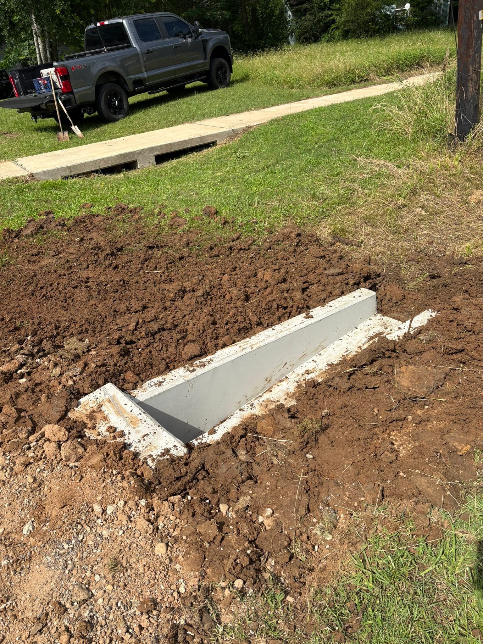 Concrete drainage culvert installed in a dirt trench beside a grassy yard and driveway, with a pickup truck in the background