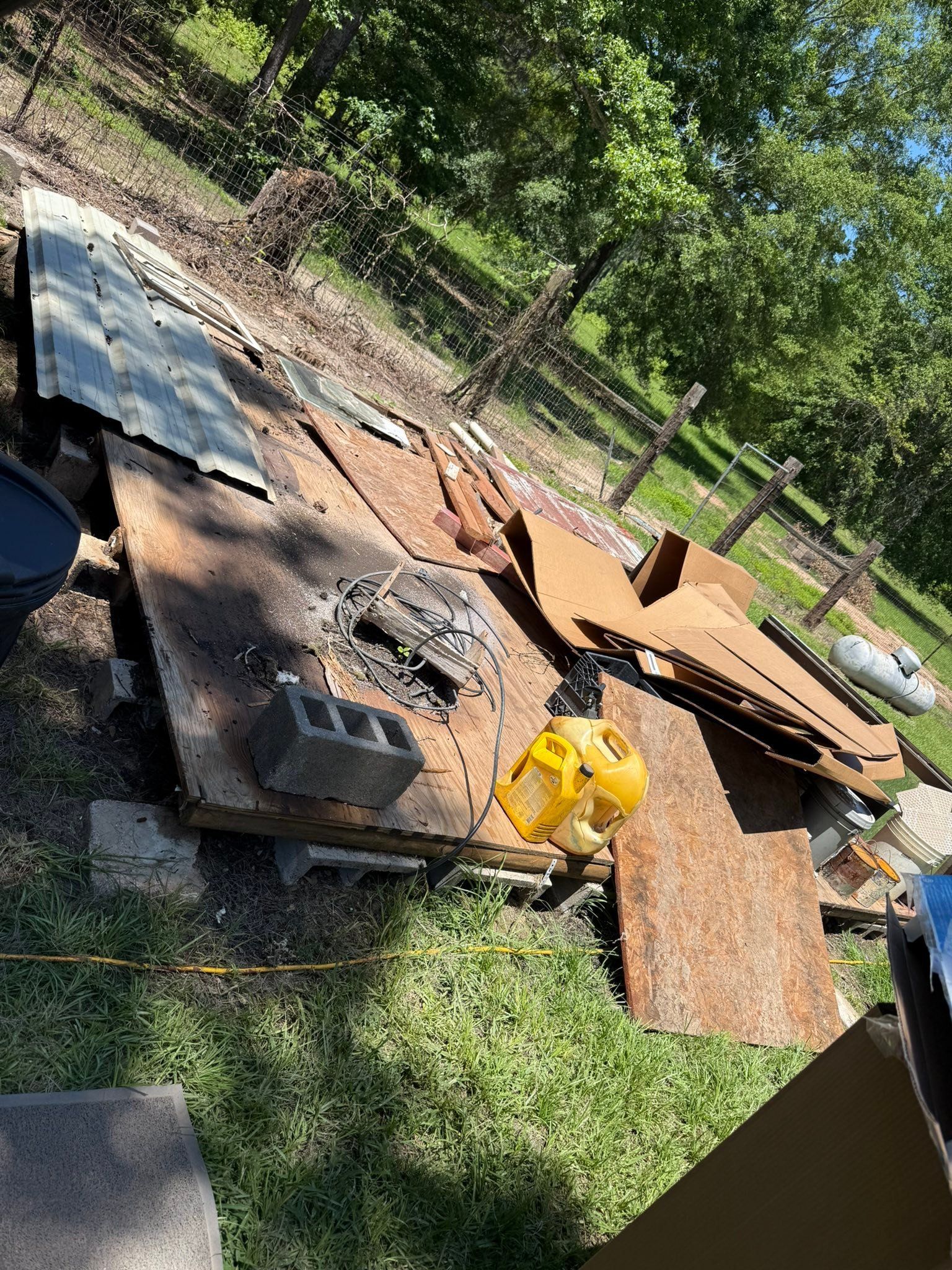 Rusty metal sheets and tools scattered around a backyard fire pit area near a fence, with grass and trees nearby.