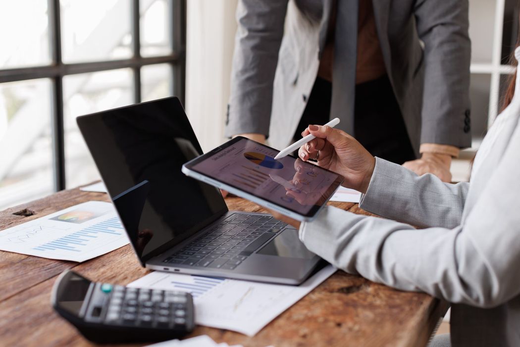 Two professionals in business attire collaborate at a wooden desk with a laptop, tablet, calculator, and financial charts.