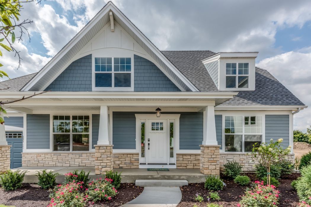 Blue and white craftsman-style house with stone accents and a gray roof. Landscaping with pink flowers.