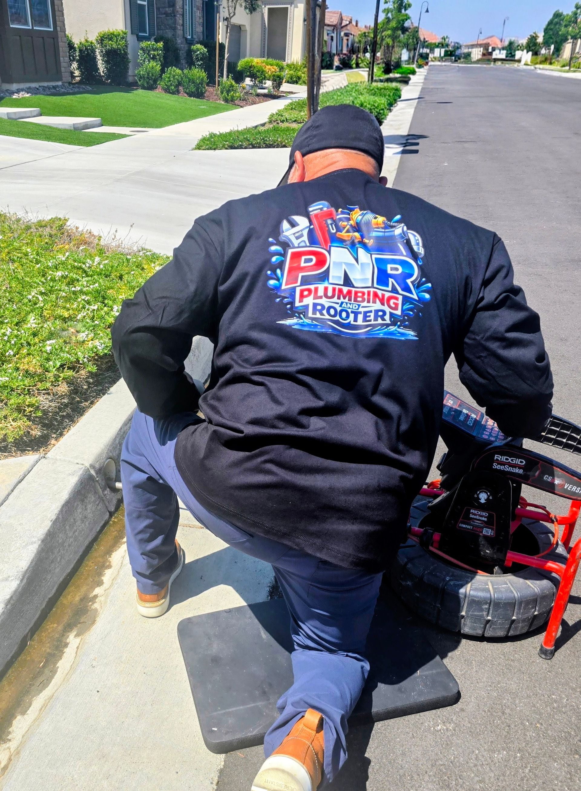Worker kneeling beside a red cart on a residential street, adjusting equipment on the curb.
