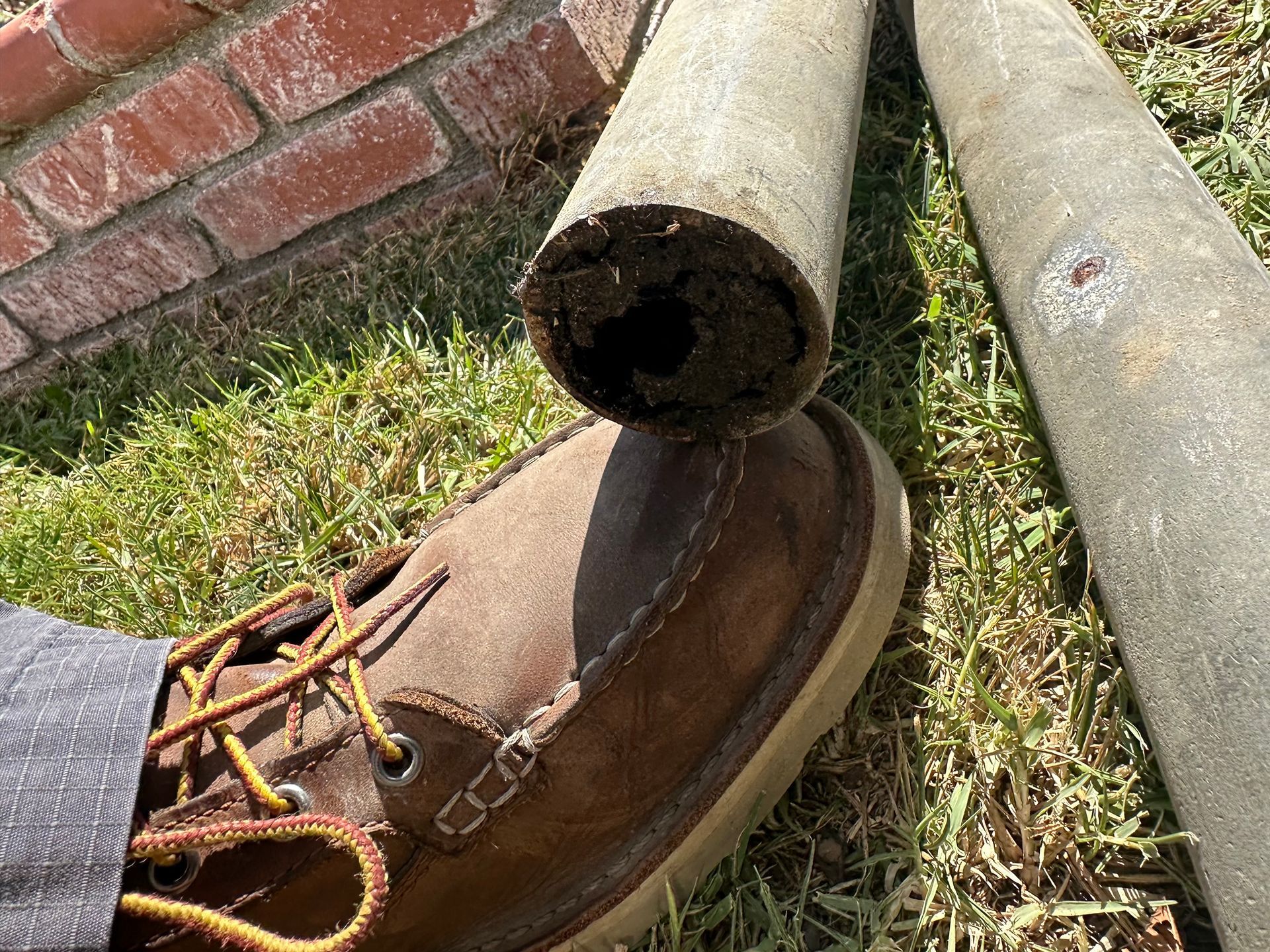 Brown hiking boot wedged under two gray pipes beside a brick wall on grass