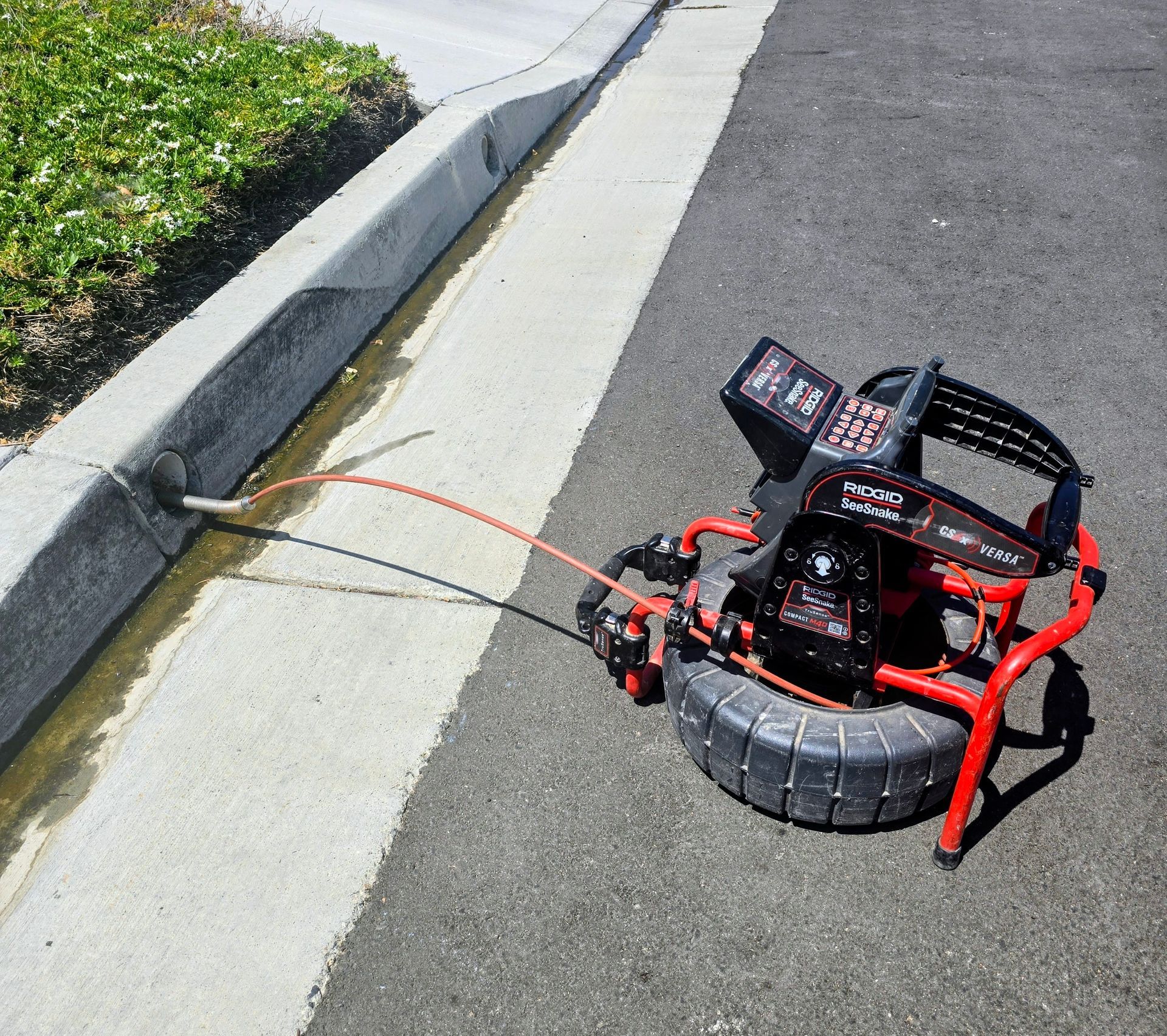 Red remote-control robot on asphalt, beside a curb and grassy verge, with wires trailing out.