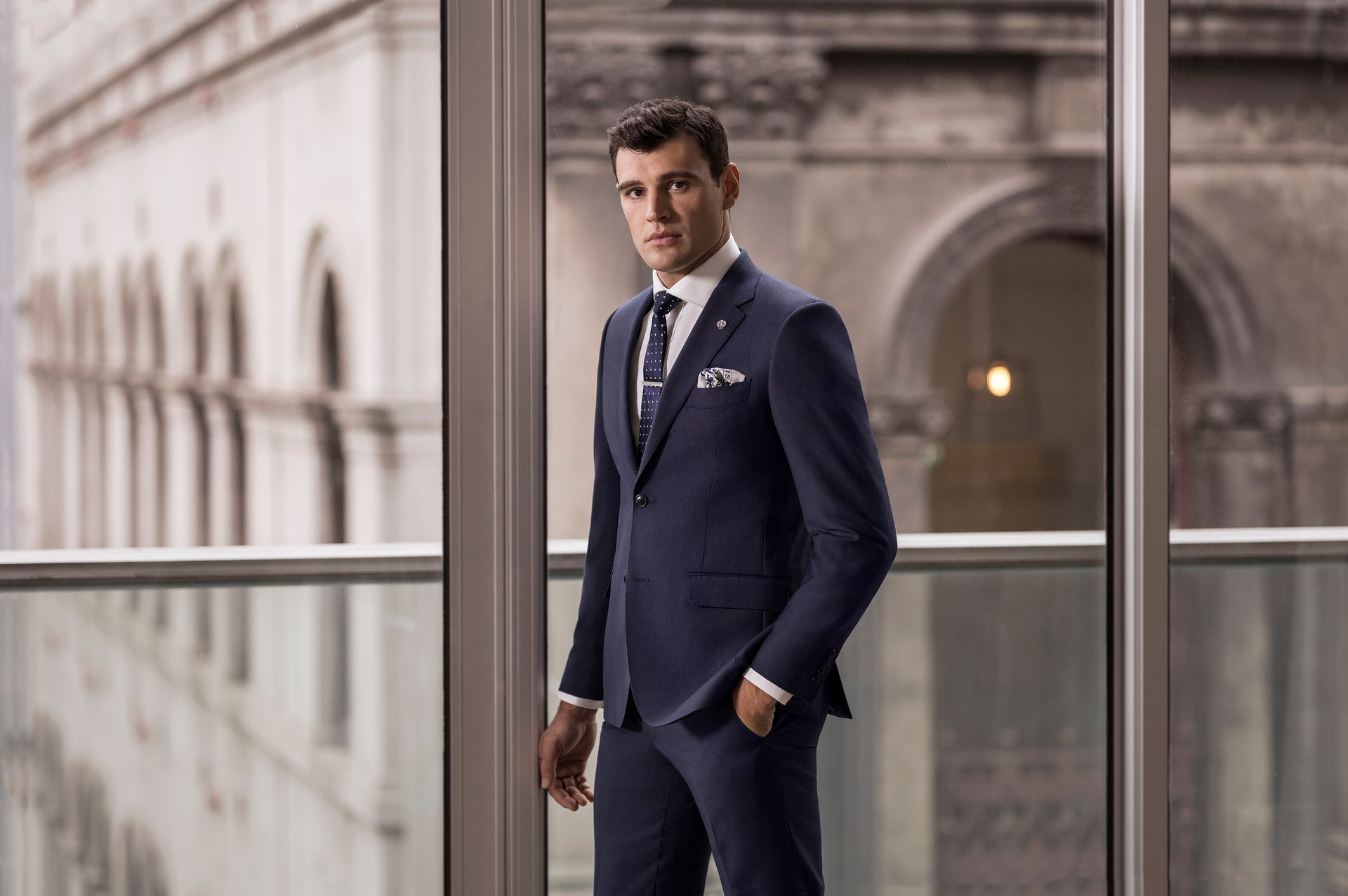 Man in a navy suit standing by a window in a modern office building