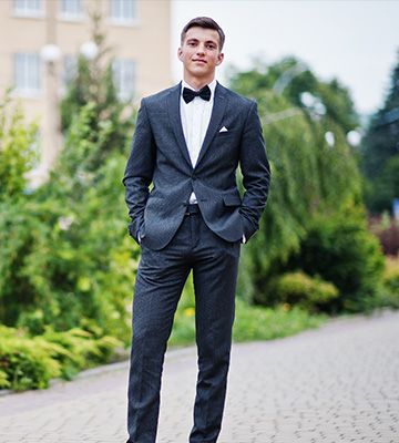 Man in a dark suit and bow tie standing on a paved path outdoors, hands in pockets.