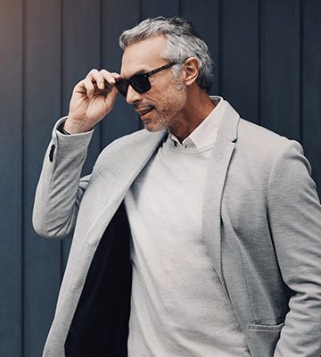 Man adjusting sunglasses outdoors, wearing a light gray blazer and white shirt against a dark wall
