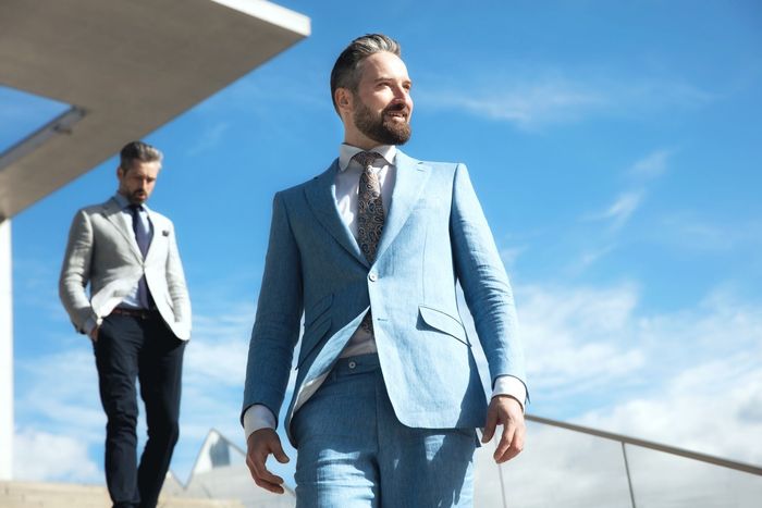 Two men in suits walking outdoors under a blue sky, one in a light blue suit in front.
