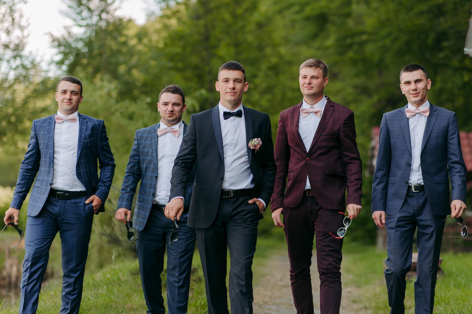 Five men in suits walking outdoors on a grassy path, two carrying jackets, with trees in the background