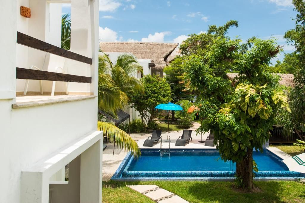 Resort courtyard with blue pool, palm trees, lounge chairs, and a turquoise umbrella under a bright sky
