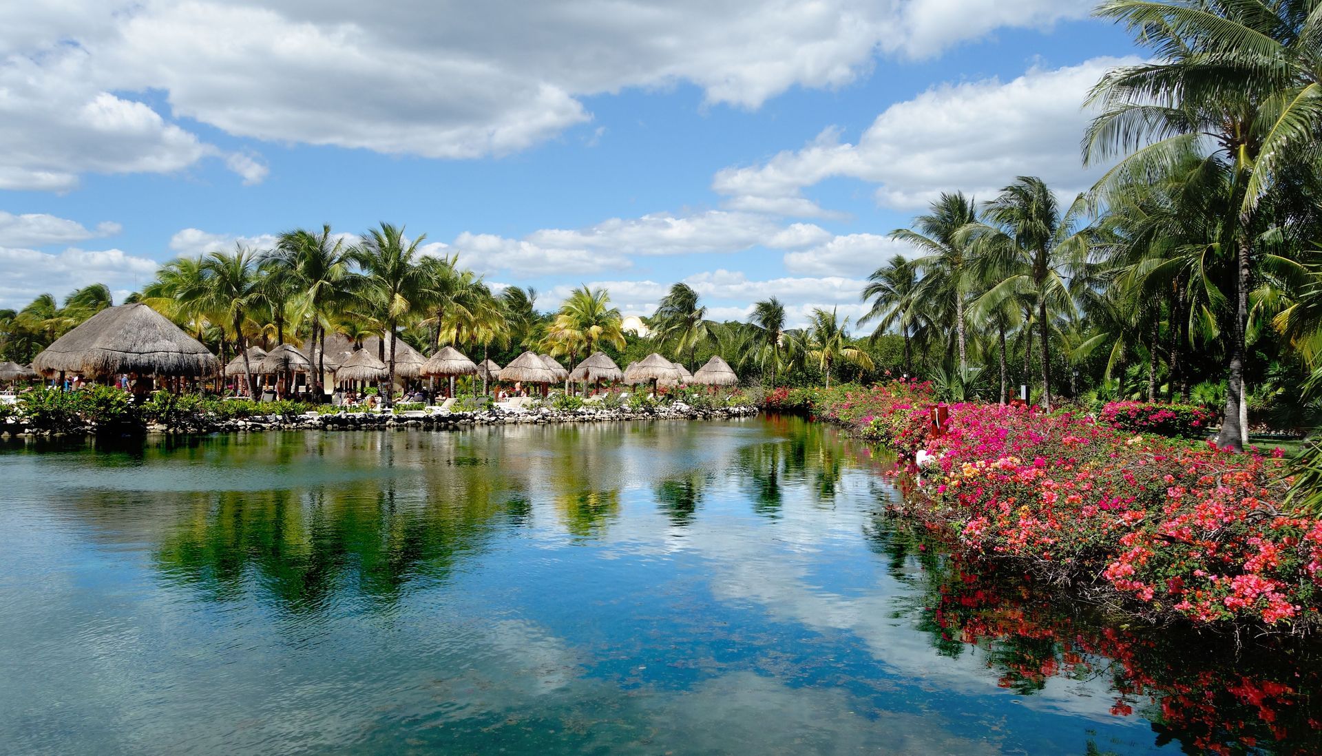 Tropical resort lagoon with thatched huts, palm trees, and pink flowers reflecting in calm water