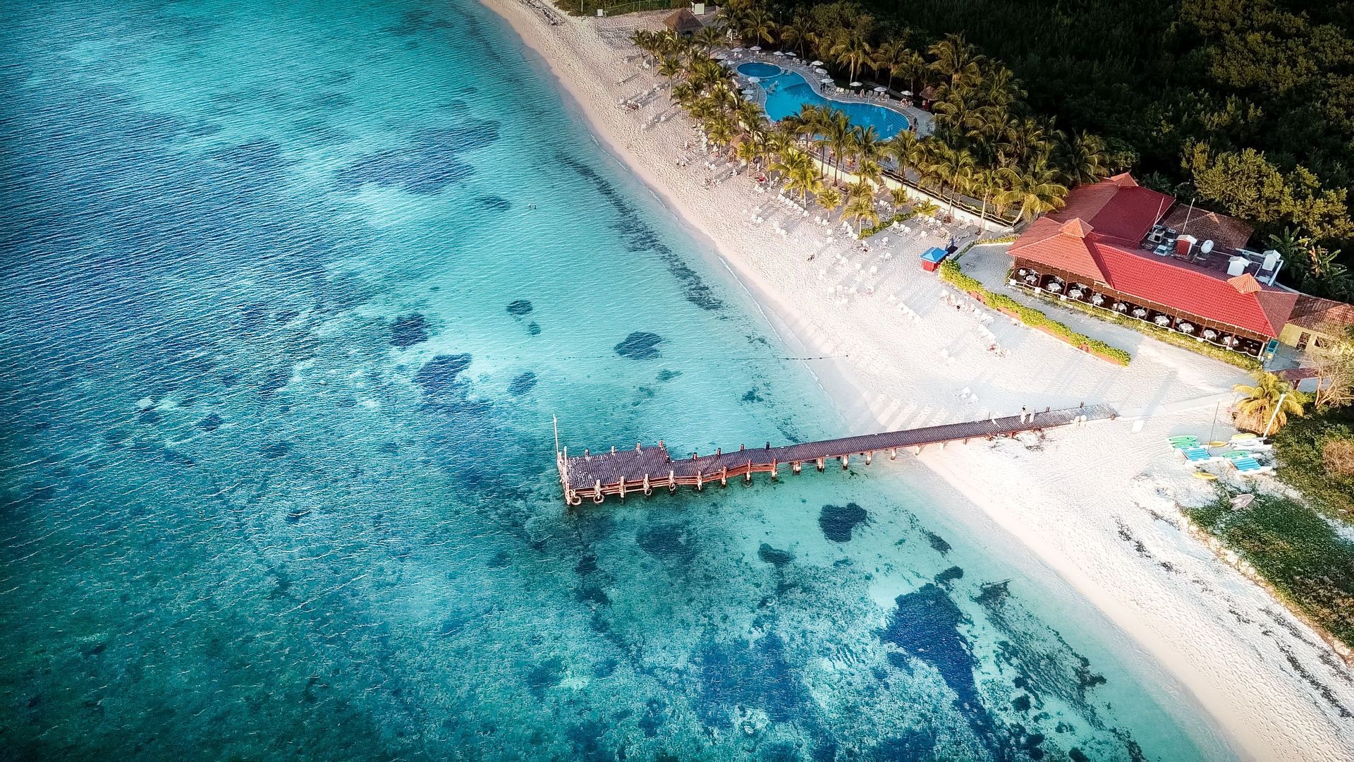 Aerial view of a tropical beach with turquoise water, white sand, a wooden pier, and a red-roofed building.