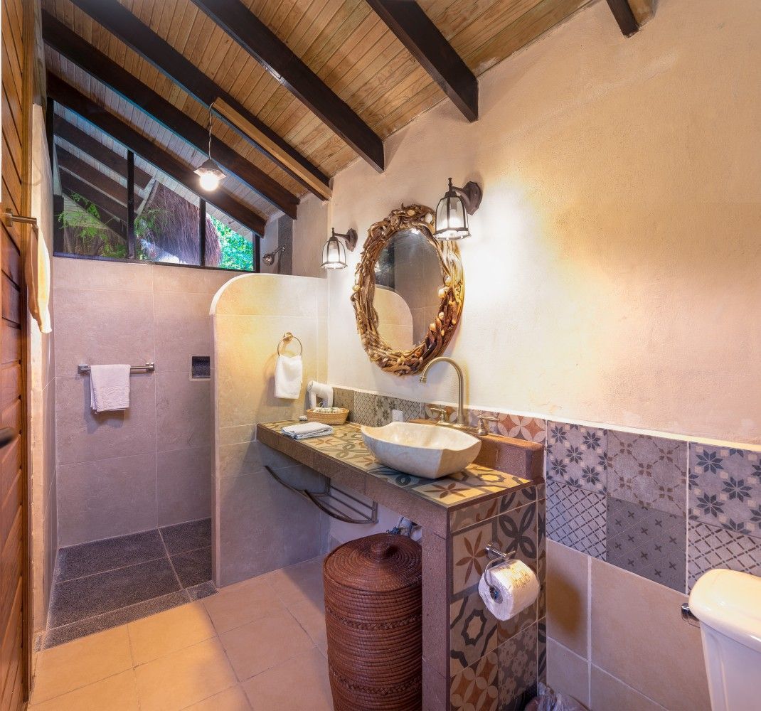 Stone-and-wood bathroom with vessel sink, ornate mirror, toilet, and open shower under a sloped ceiling