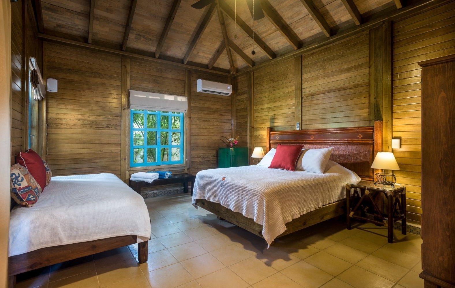 Rustic bedroom with two beds, wood-paneled walls, tiled floor, and a window letting in daylight