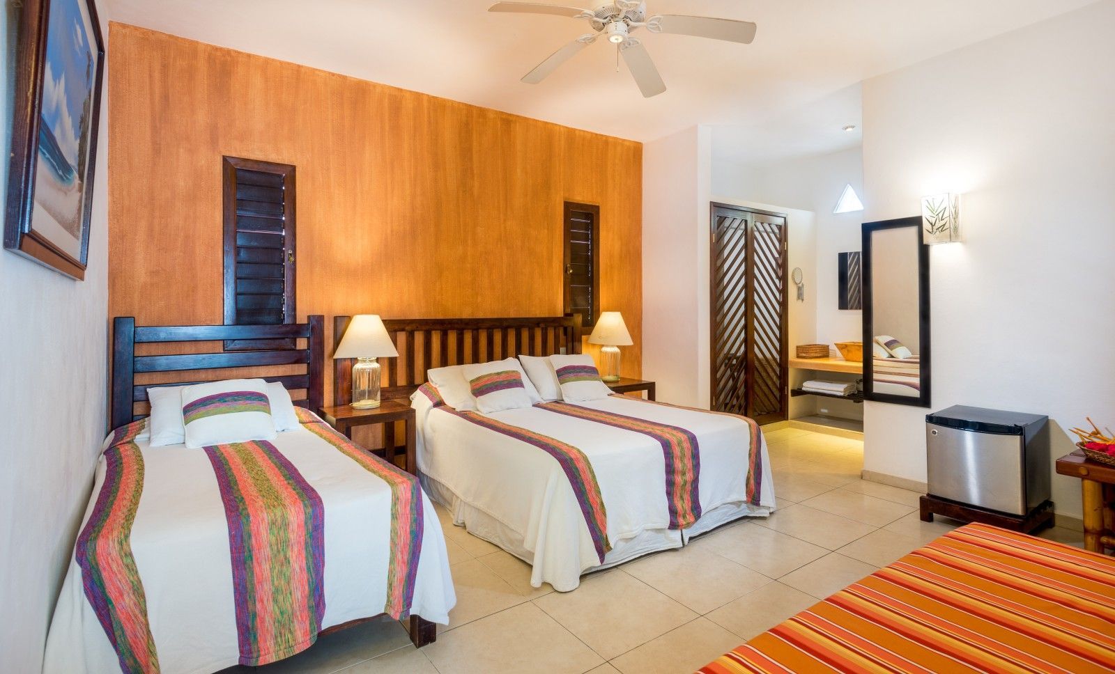 Bright hotel room with two twin beds, wood accent wall, lamp, dresser, and tiled floor