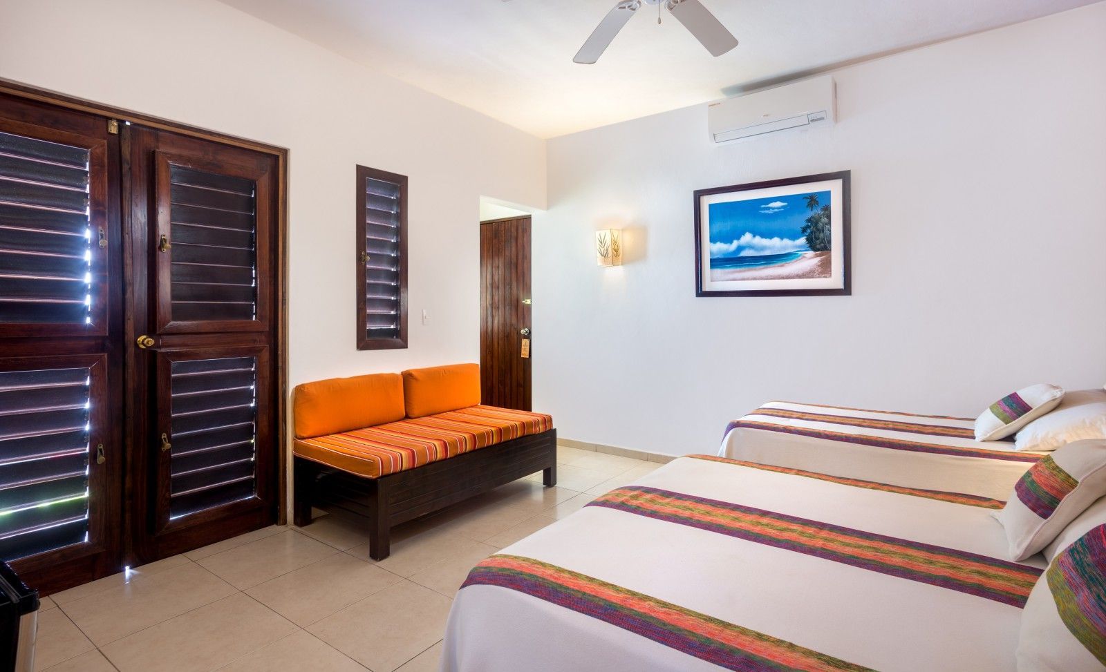 Bright bedroom with twin beds, orange bench, ceiling fan, window shutters, and wall art
