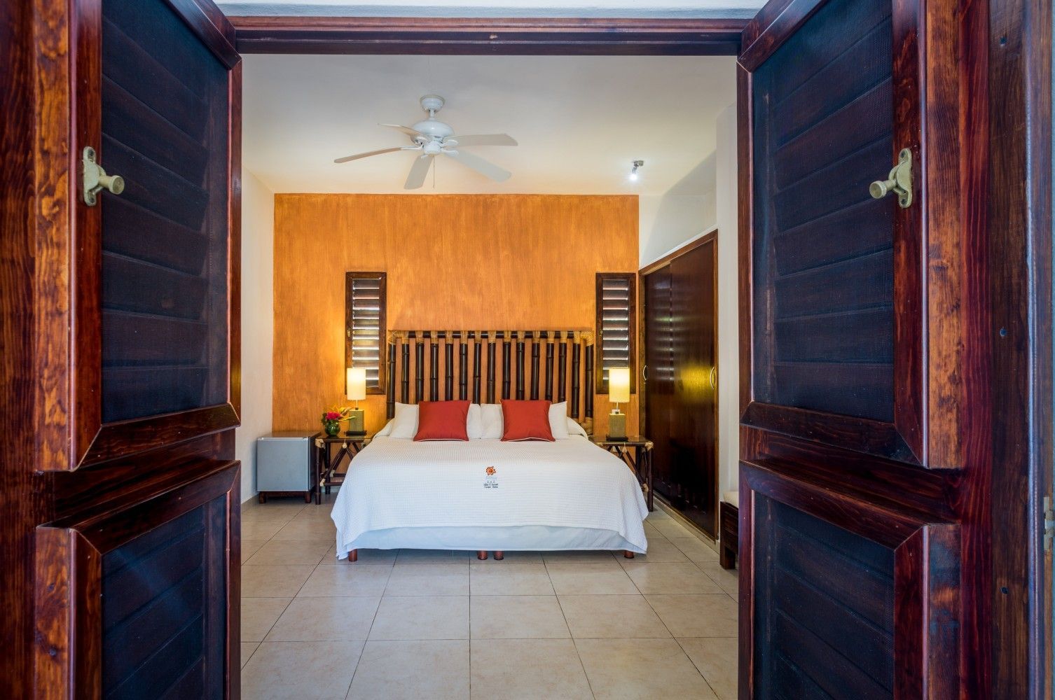 Bedroom viewed through open doorway with white bed, orange accent wall, and dark wood shutters