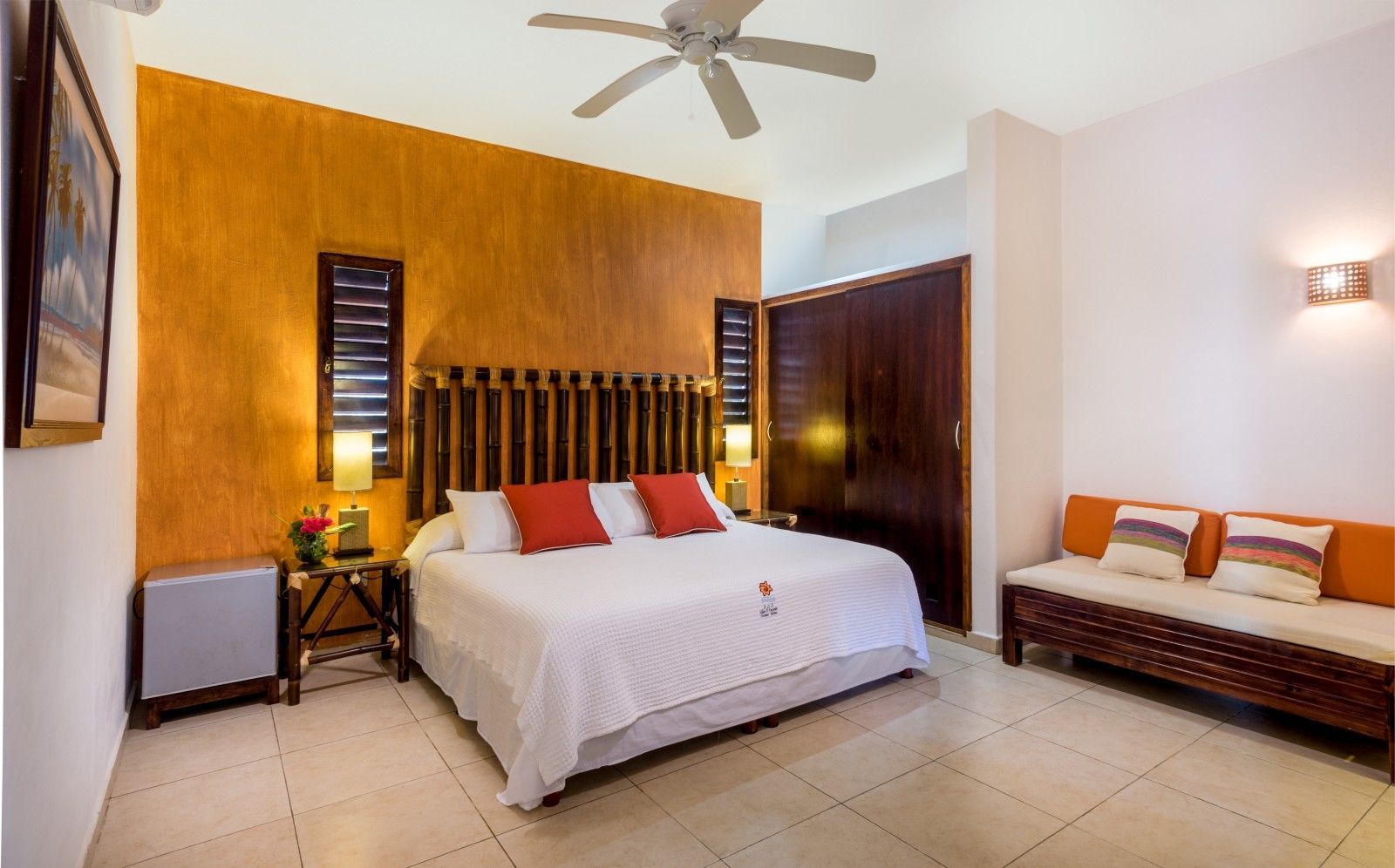 Cozy bedroom with white bed, orange accent wall, wooden headboard, and a bench with cushions.