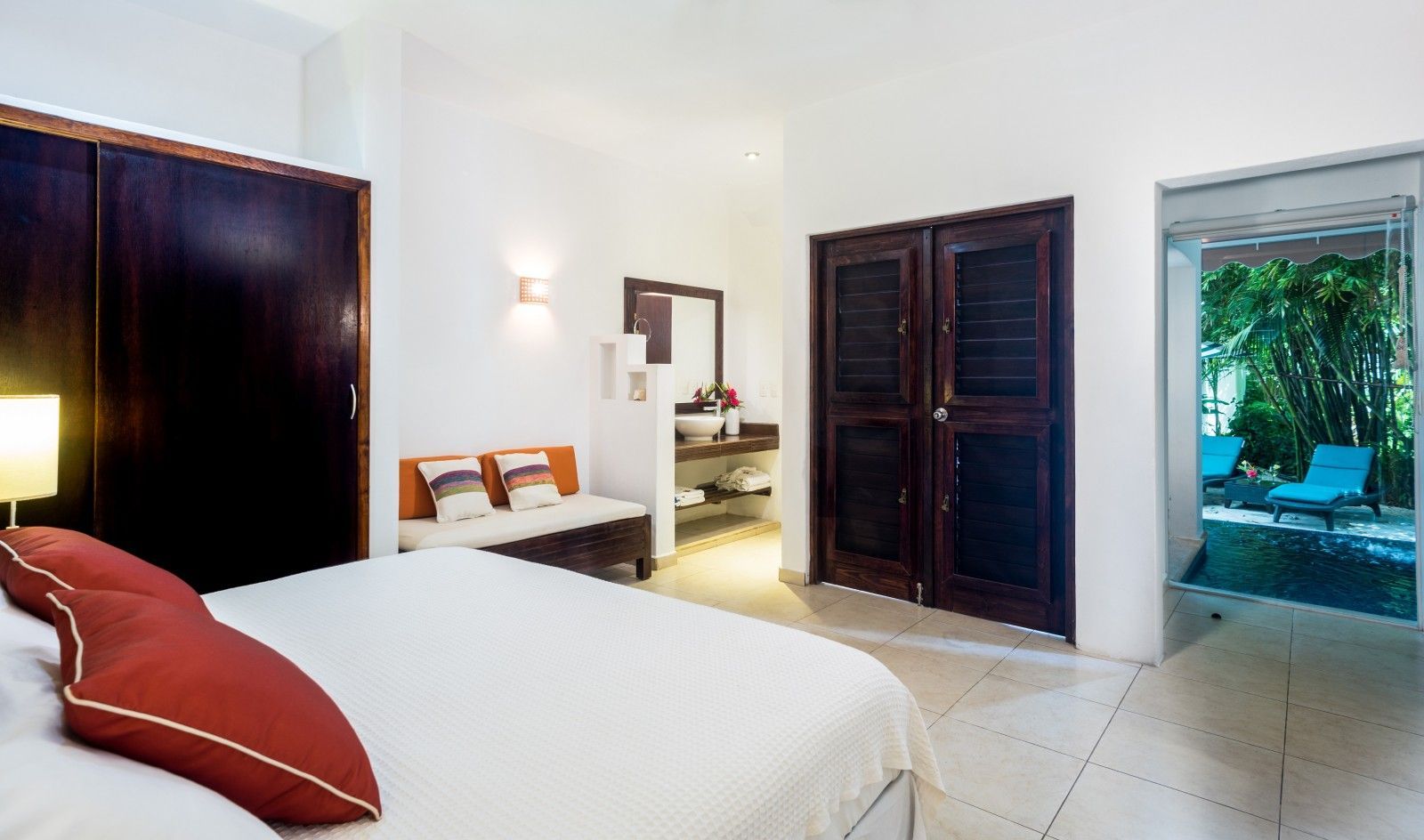 Bright bedroom with white bed, dark wardrobes, and open door to a tropical patio pool.