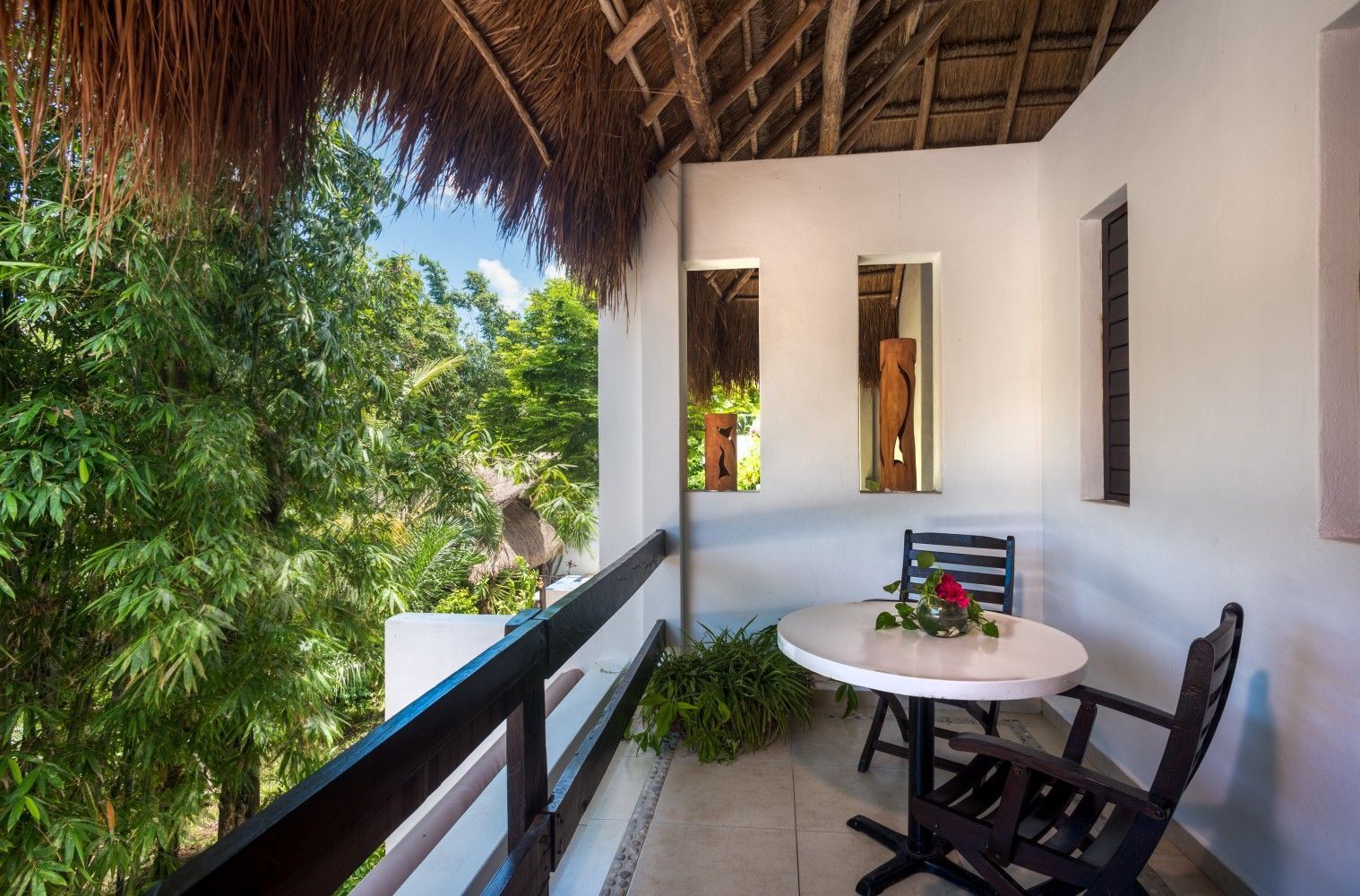 Tropical balcony with white walls, thatched roof, round table, chairs, and lush greenery beyond the railing
