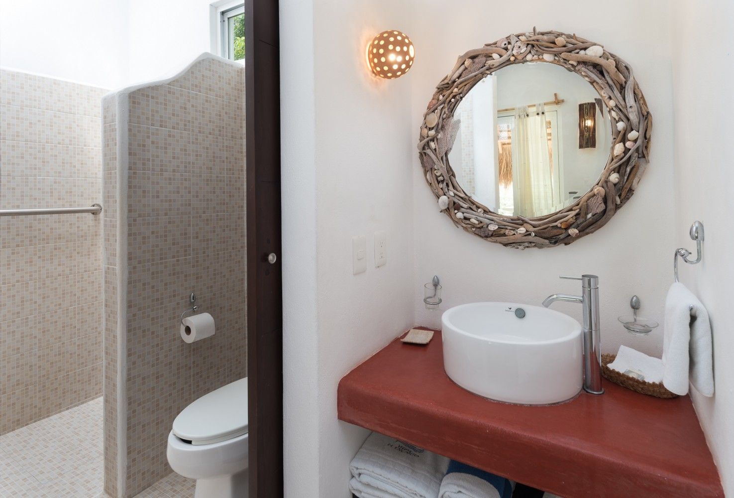 Small bathroom with a toilet stall, vessel sink on a red counter, and round mirror on the wall.