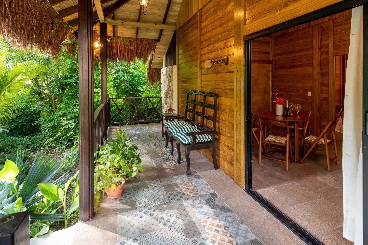 Covered tropical porch with wooden walls, stone floor, and a bench beside an open doorway.