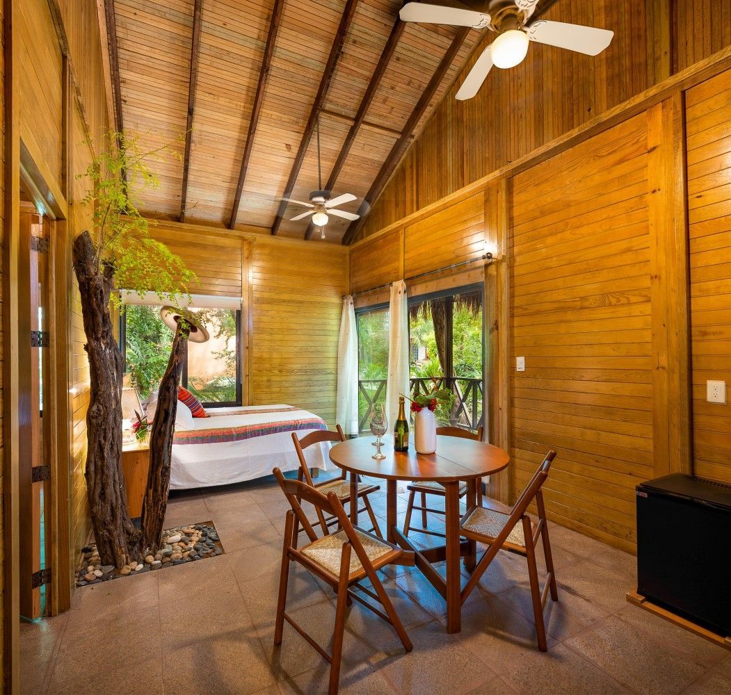 Cozy wooden bedroom with a bed, round table and chairs, ceiling fans, and tropical greenery outside the windows