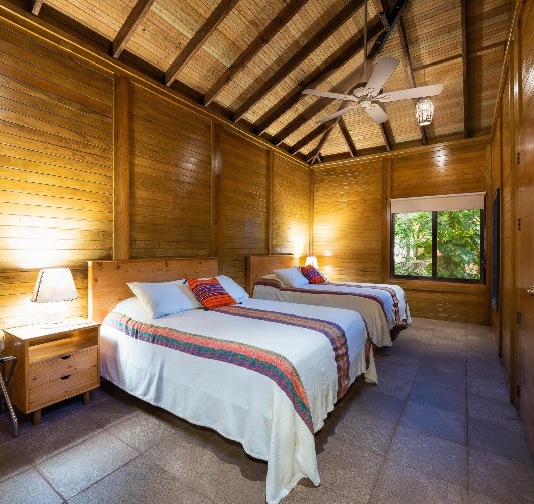 Cozy wooden bedroom with two twin beds, warm lamps, and a ceiling fan under a thatched roof