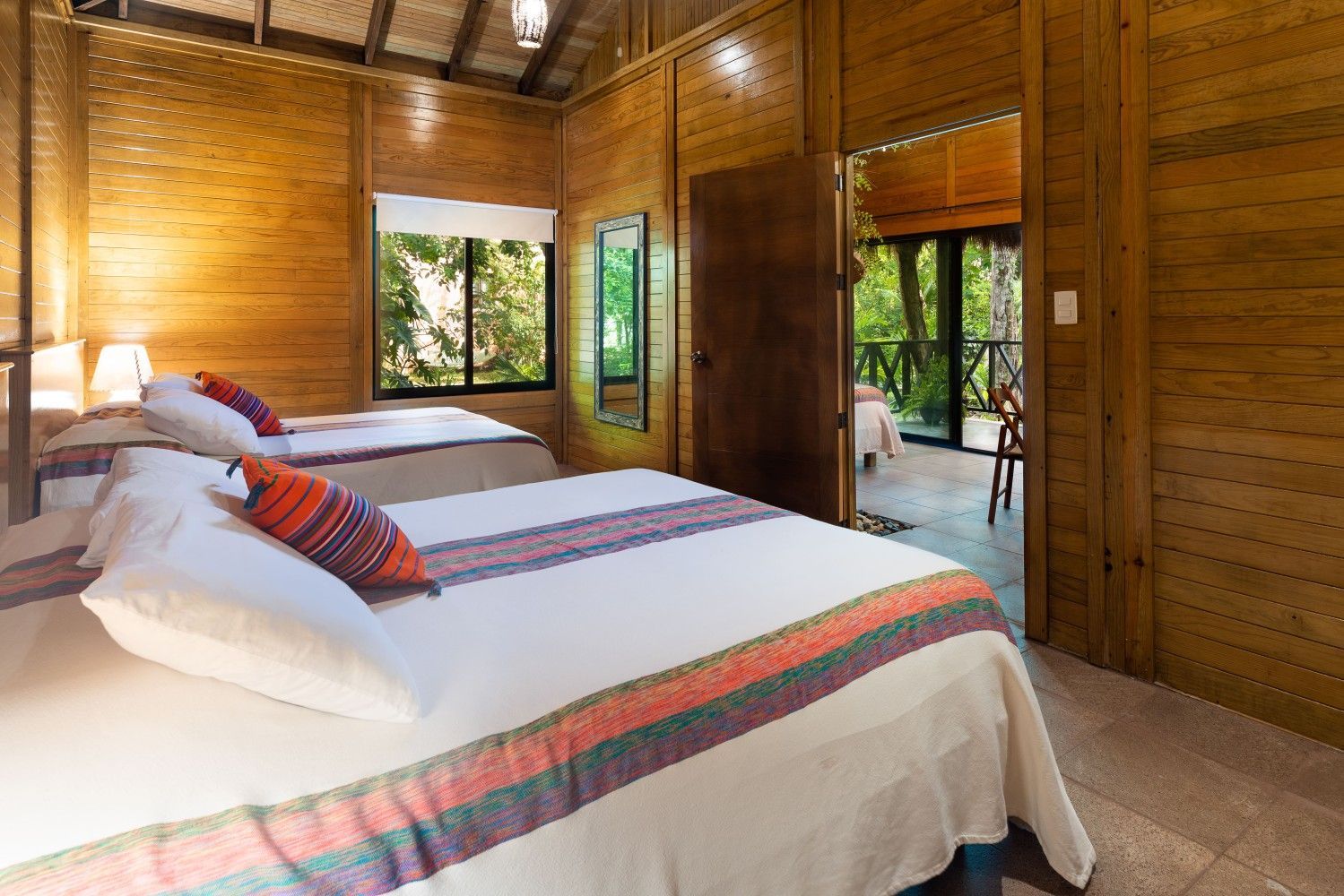 Bright wooden bedroom with a white bed, striped pillows, and open doors to a leafy outdoor view