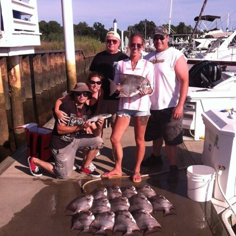 Five people posing with a large catch of fish on a dock beside boats and water.