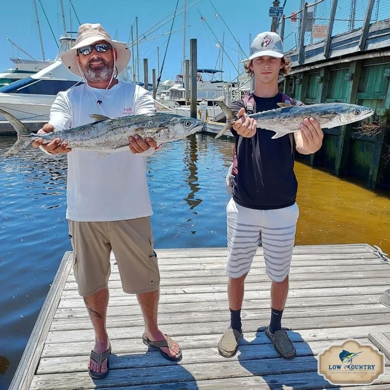 Two people on a dock holding large fish, with boats and blue water in the marina behind them.