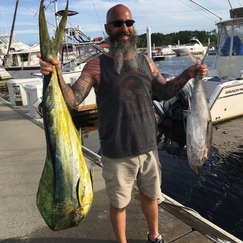 Man holding two large fish on a dock beside boats, smiling in sunglasses