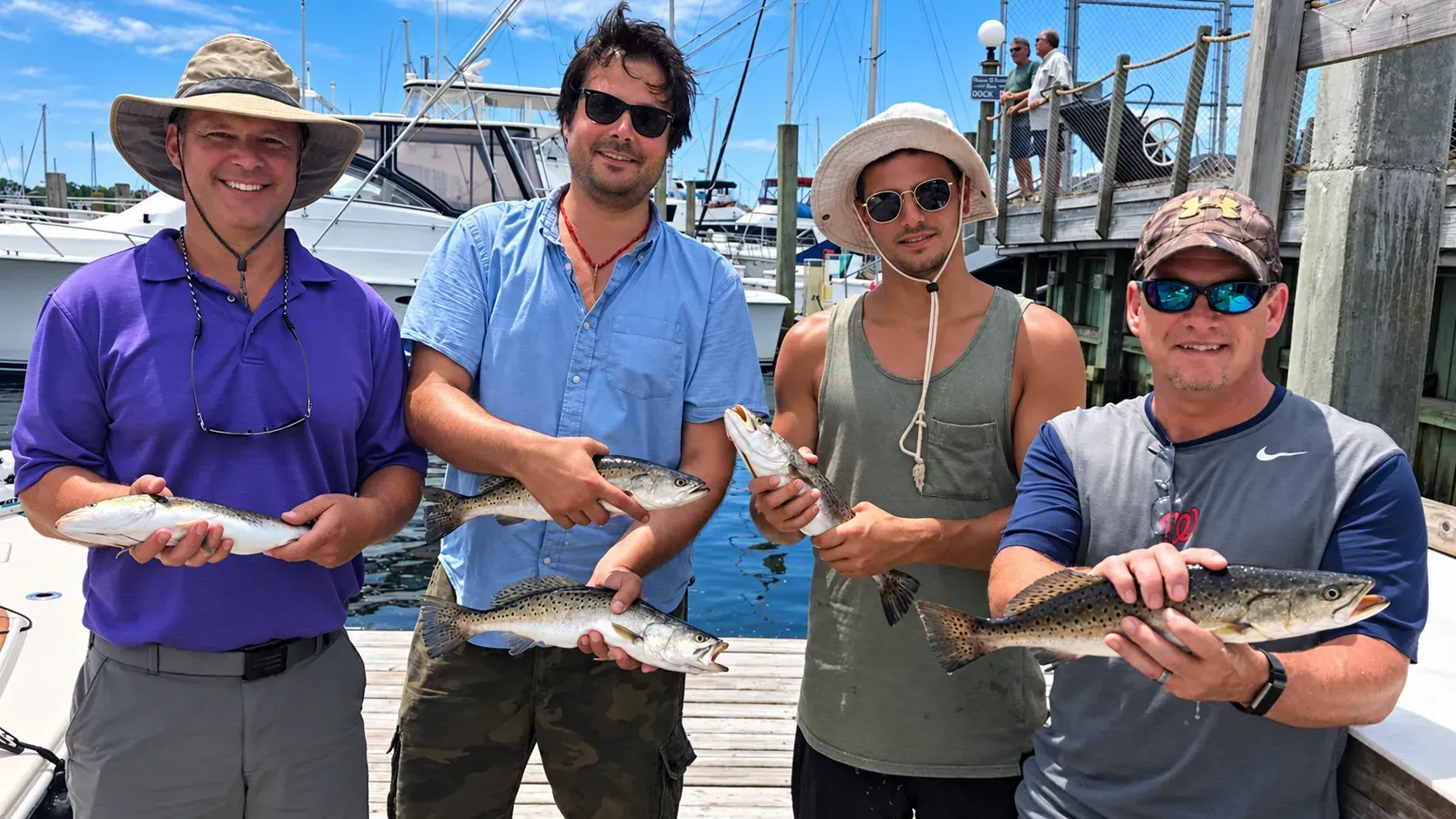 Four people on a dock holding fresh fish, with boats and a marina in the background.