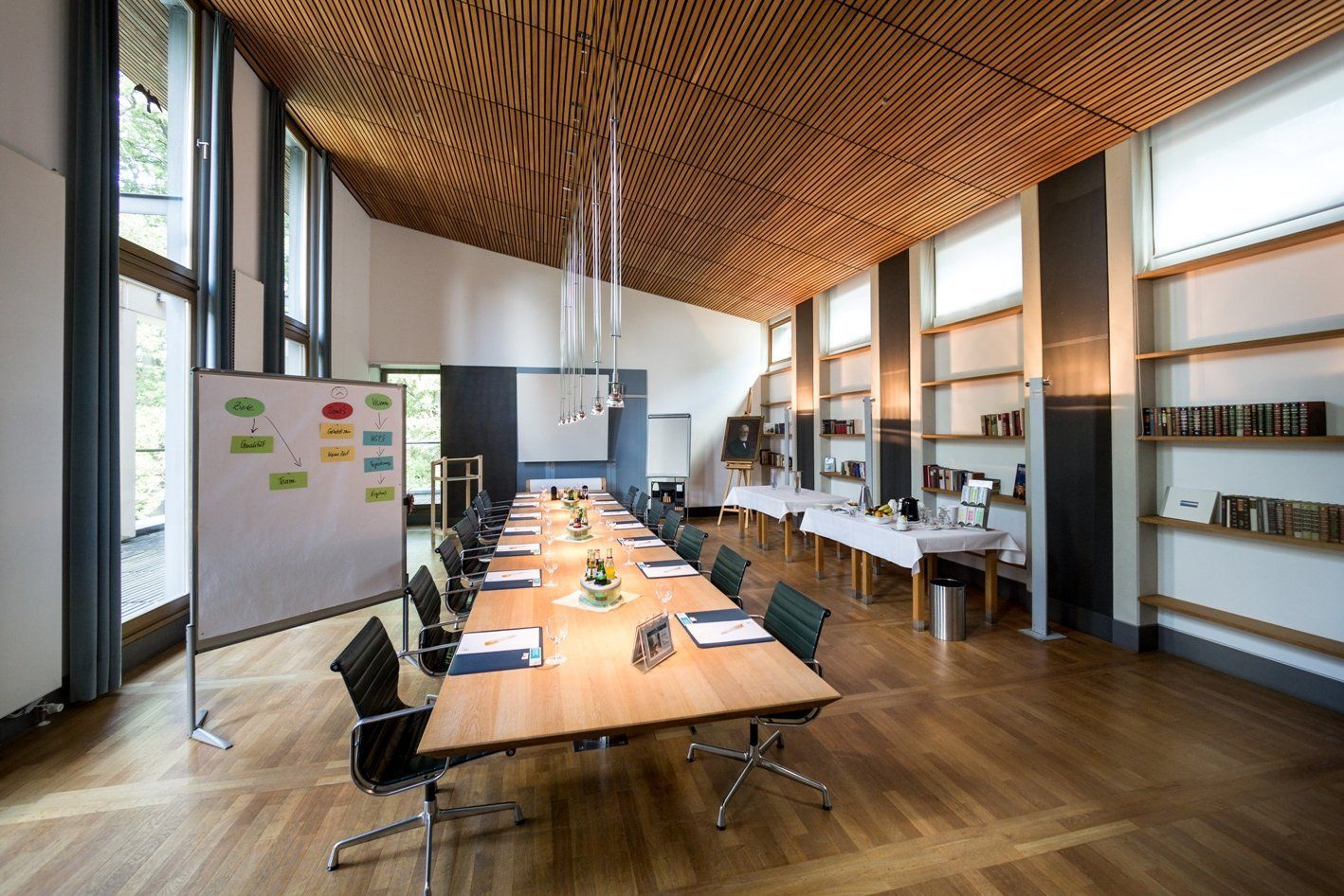 Konferenzraum mit langem Tisch, Stühlen, Whiteboard, Holzdecke und Fenstern.
