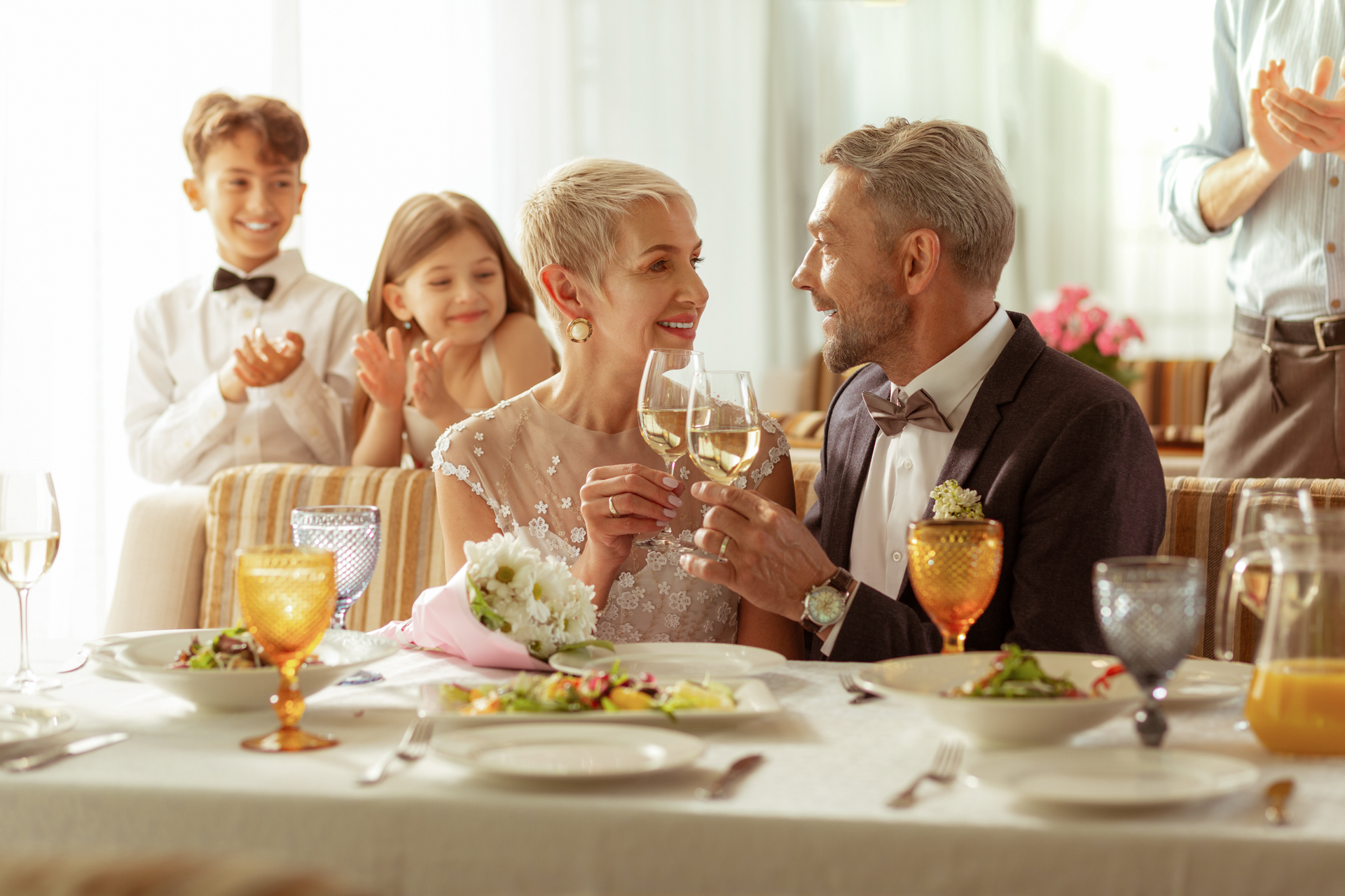 Älteres Paar stößt bei einem Hochzeitsempfang an, lächelt sich an, Kinder und Gäste klatschen.