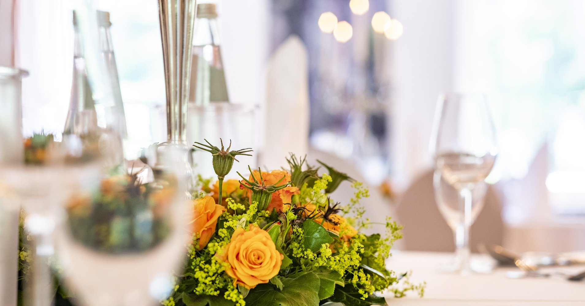 Tafelaufsatz aus orangefarbenen Rosen und Grünpflanzen auf einer weißen Tischdecke, mit verschwommenem Glasgeschirr und Wasserflaschen im Hintergrund.