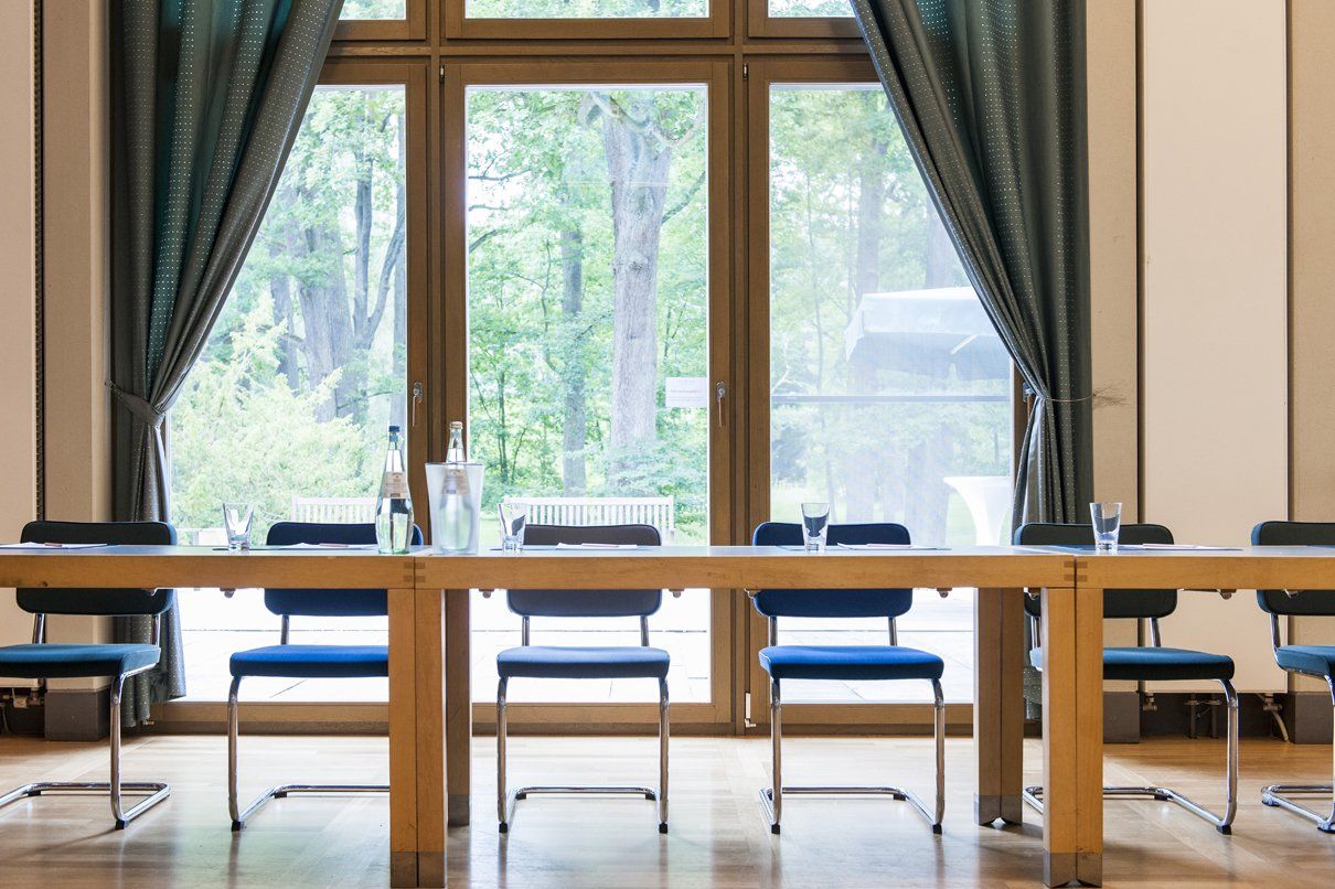 Langer Holztisch mit blauen Stühlen vor einem großen Fenster mit grünen Vorhängen und Blick auf Bäume.