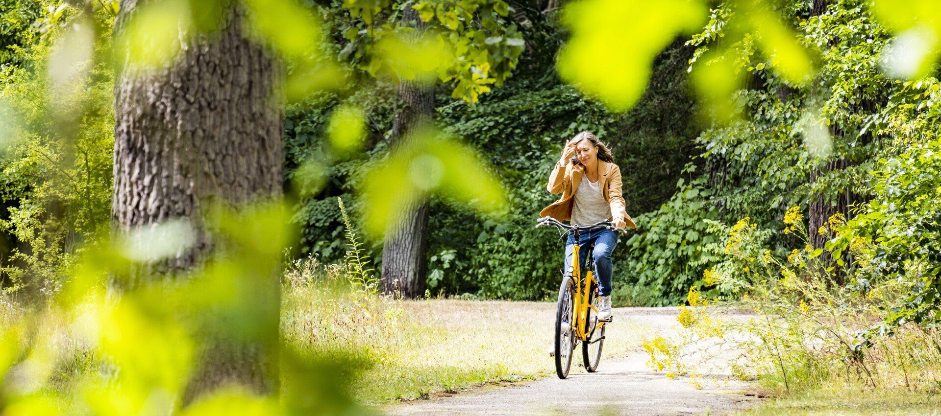 Frau fährt auf einem gelben Fahrrad auf einem Weg durch einen sonnenbeschienenen Wald und hebt ihre Hand.
