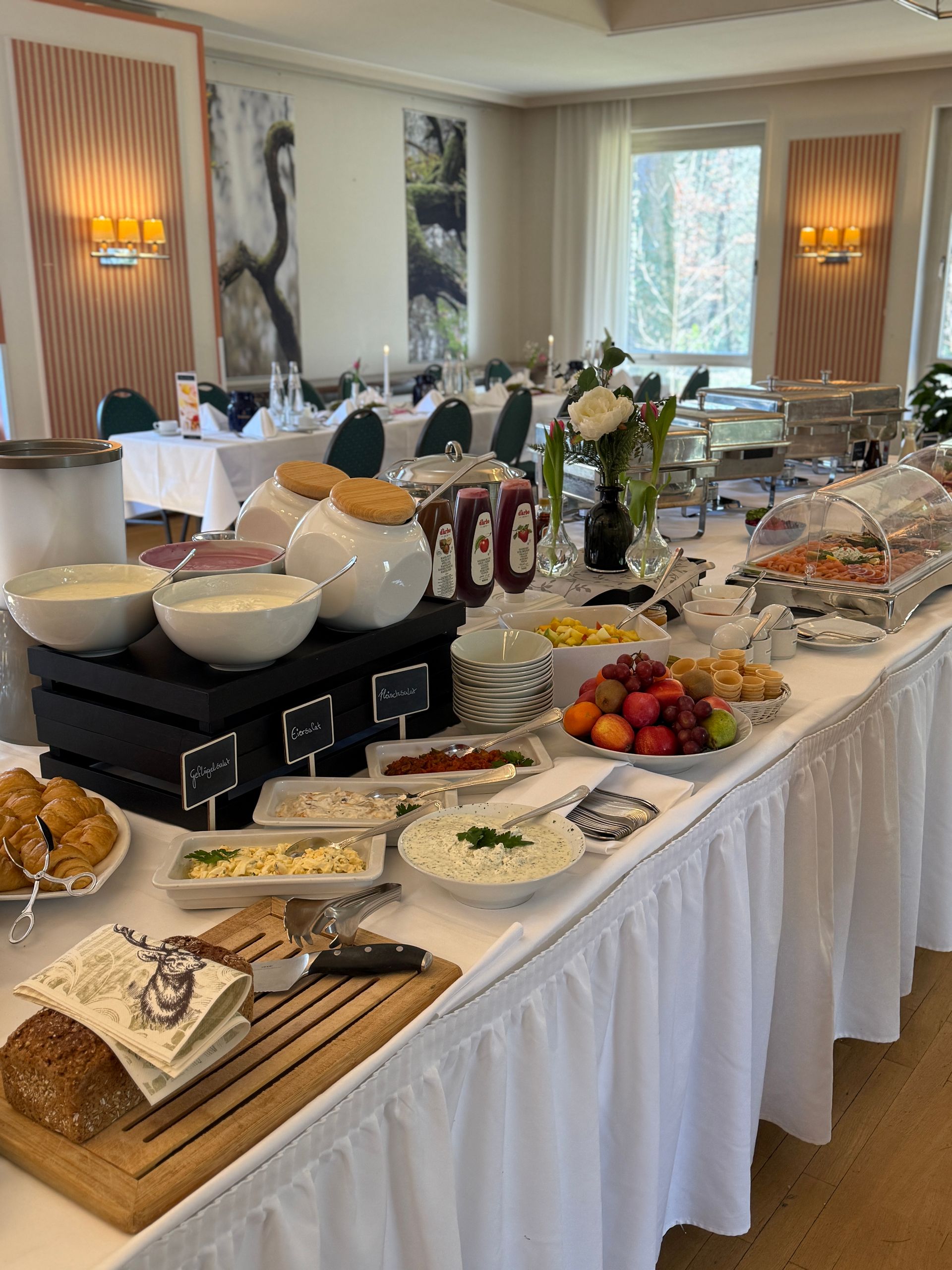 Buffettisch in einem gut beleuchteten Raum mit verschiedenen Lebensmitteln: Joghurt, Obst, Brot, Gebäck.