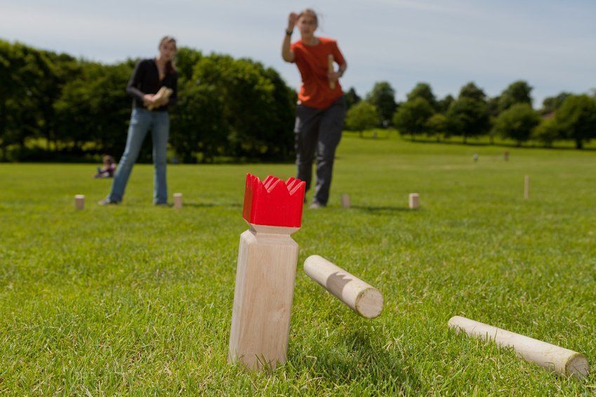 Zwei Personen spielen ein Rasenspiel; einer wirft einen Holzscheit auf eine Königsfigur. Grünes Gras, sonniger Tag.
