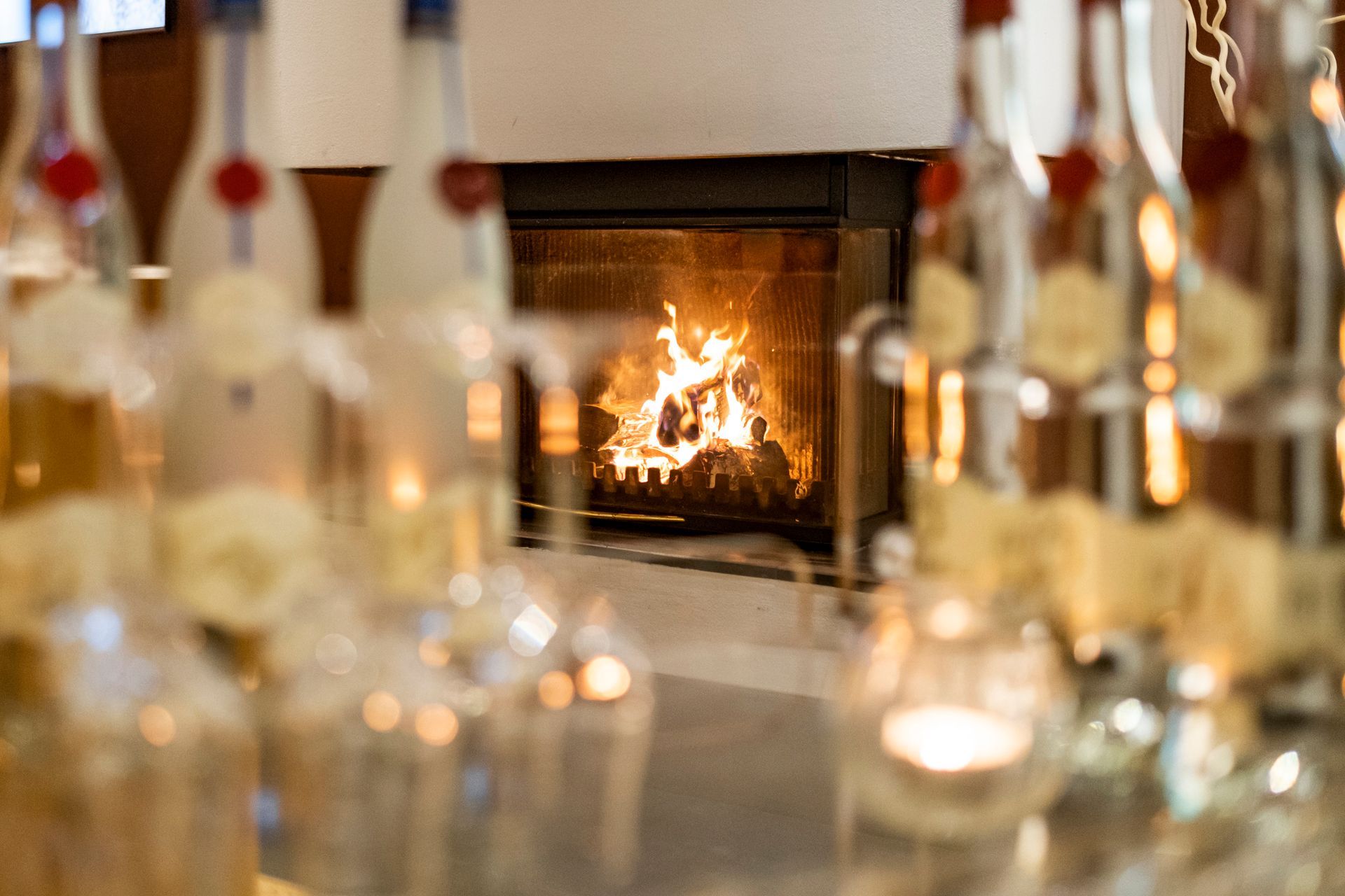Kamin mit Flammen, durch verschwommene Flaschen und Kerzenhalter betrachtet. Warmes Leuchten, gemütliche Atmosphäre.