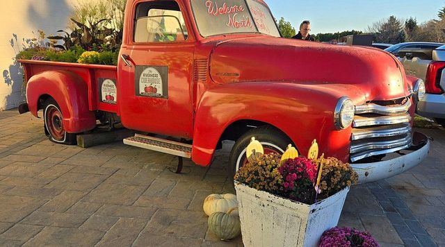 A red truck is parked on the side of the road next to flowers and pumpkins.