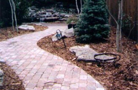 Brick pathway winding through a garden, bordered by mulch, trees, and decorative elements.