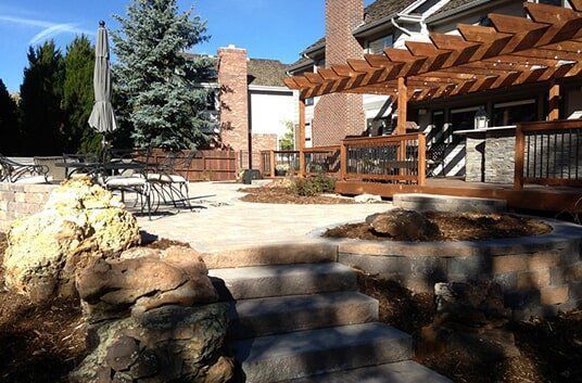 Stone patio with stairs, wood pergola, and chairs on a sunny day.