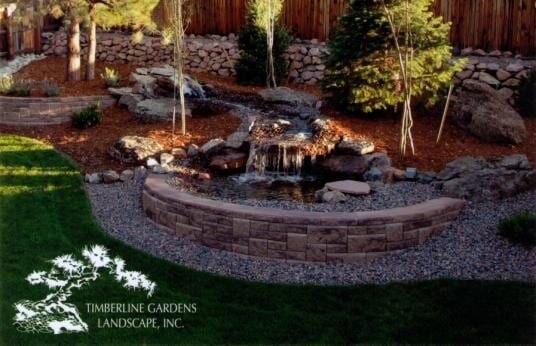 A landscaped garden with a waterfall, pond, and stone wall; green grass in foreground.