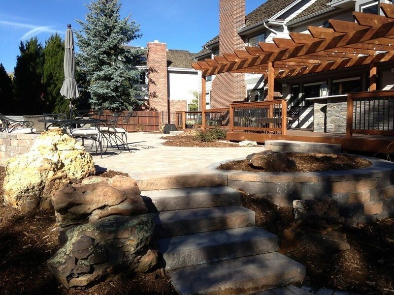 Stone patio with steps, wooden deck with pergola, and landscaping in a sunny outdoor setting.